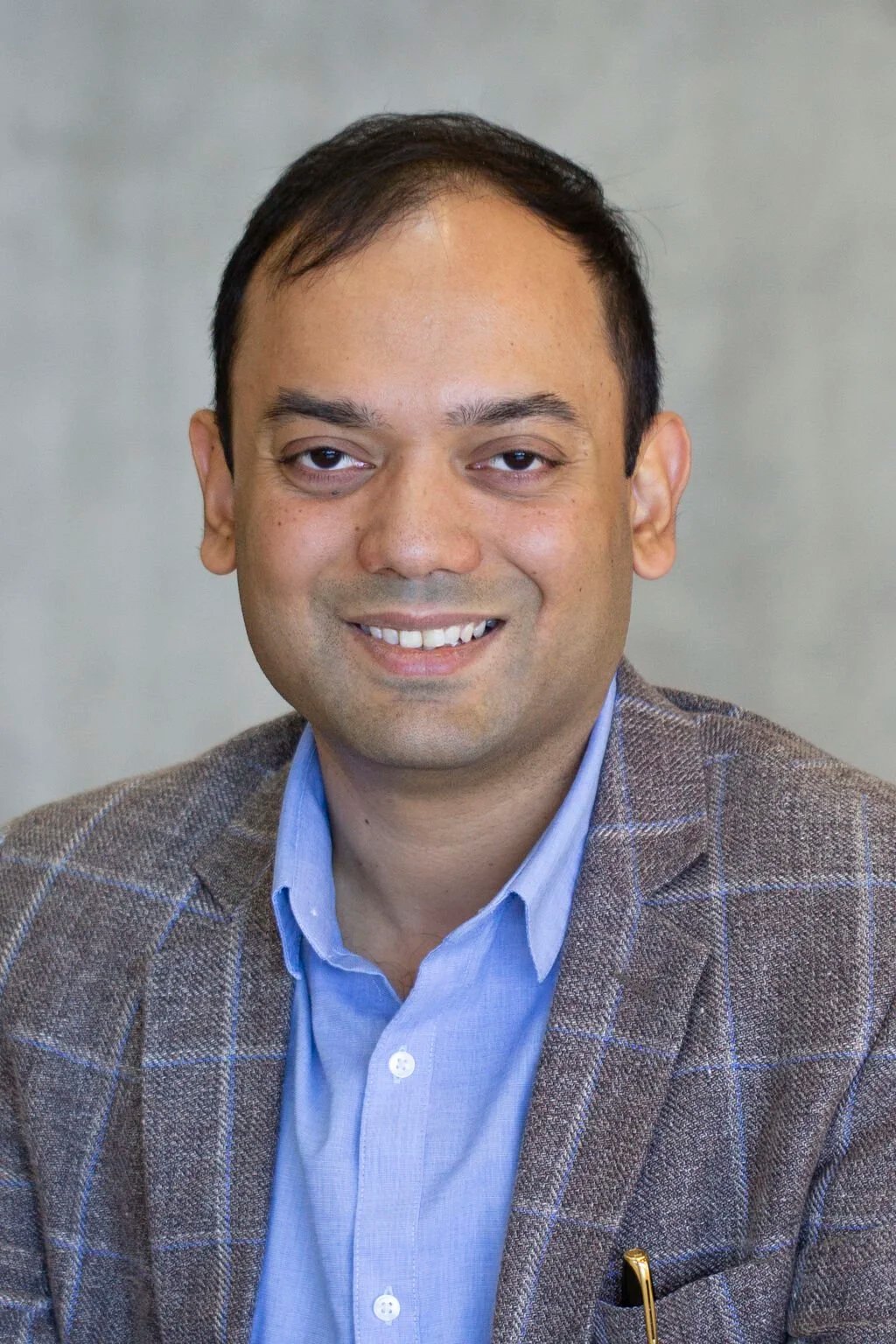 A man with short dark hair and medium skin tone, smiling, wearing a light blue shirt and a brown checkered blazer, against a plain background.