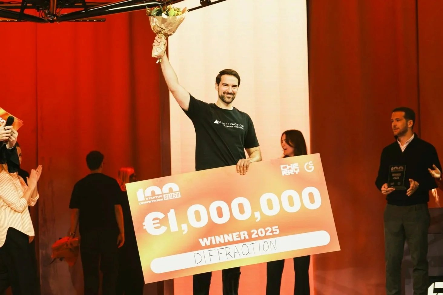 Man in black shirt holding a box, standing in front of a digital display, with green chairs behind him.