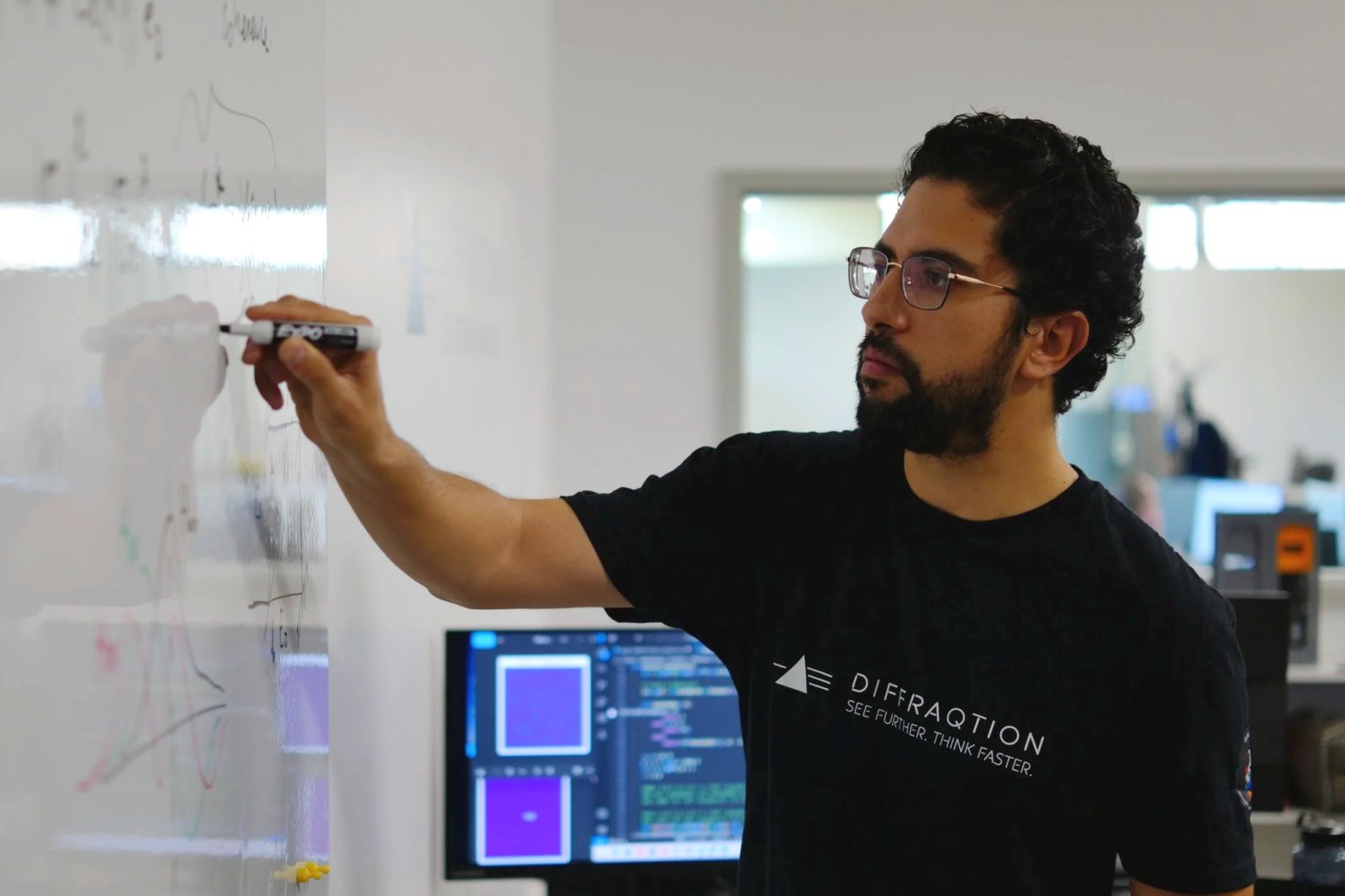 A man with dark curly hair, glasses, and a beard writes on a whiteboard with a marker. He wears a black T-shirt with the text 'DIFFRACTION SEE FURTHER. THINK FASTER' and is in a modern office or lab environment with computer screens in the background.
