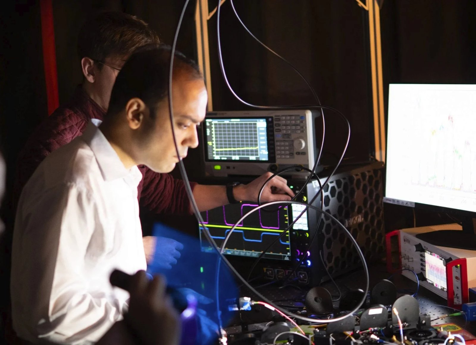 Three men working with electronic equipment, oscilloscopes, and monitors in a dark room, possibly conducting experiments or tests.