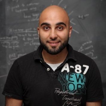Young man with a beard and shaved head posing in front of a chalkboard with writing, wearing a black polo shirt with patches and text.